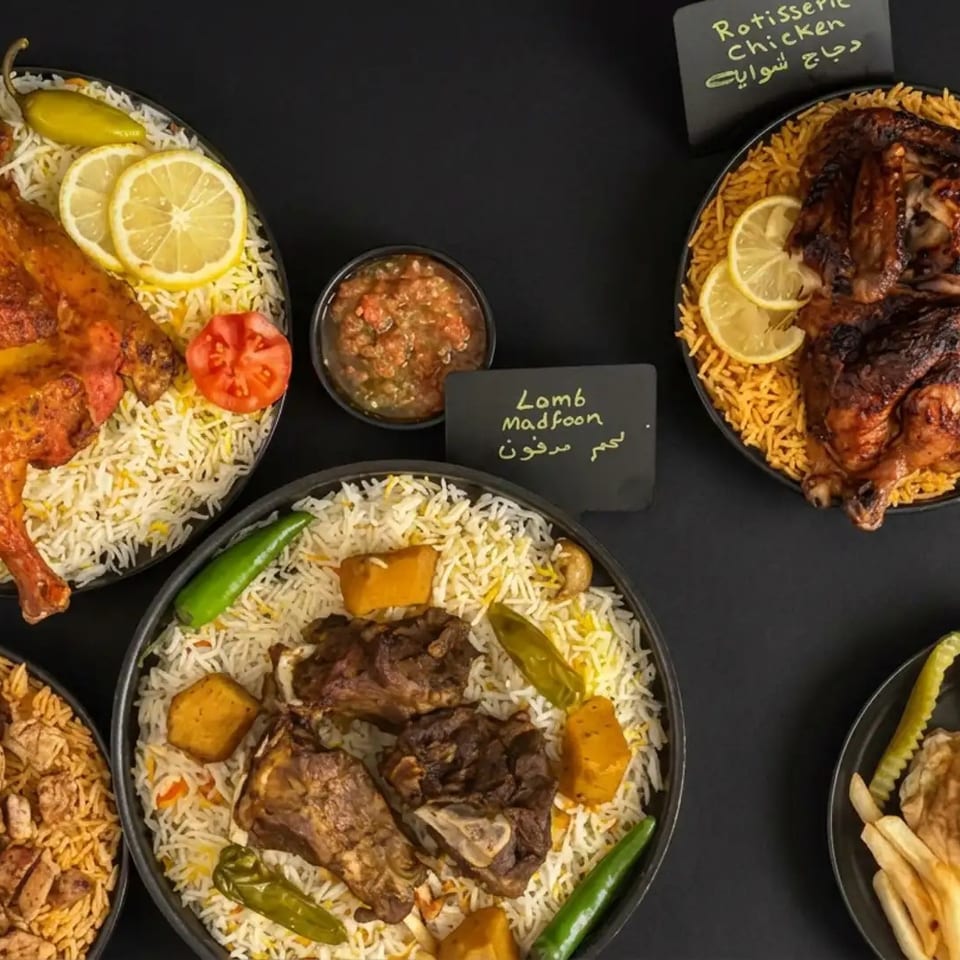 Assorted Middle Eastern dishes arranged on black plates, including roast chicken, lamb, shawarma, grilled chicken, rice, lemons, and pickles.