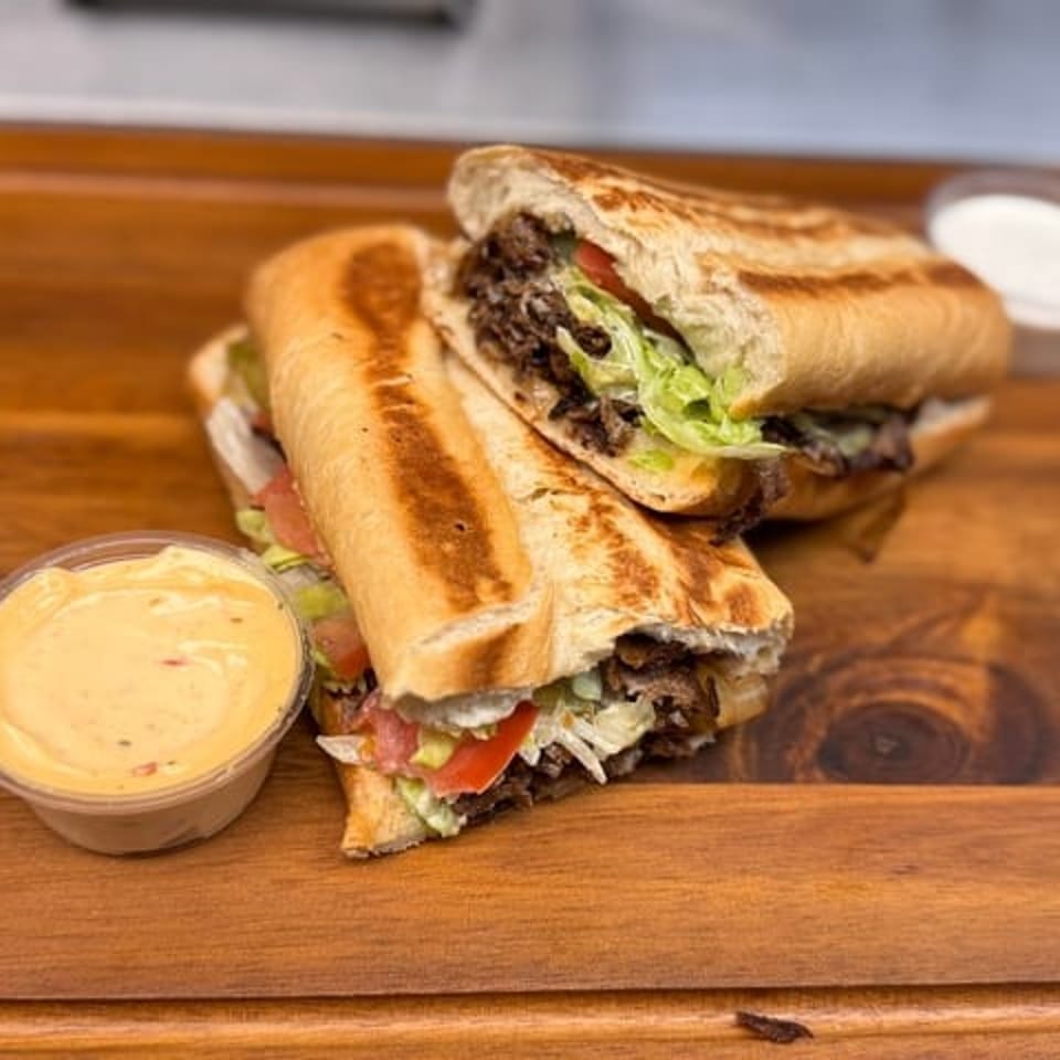 Grilled beef sandwiches with lettuce, tomato, and sauce served on a wooden board with dipping cups.
